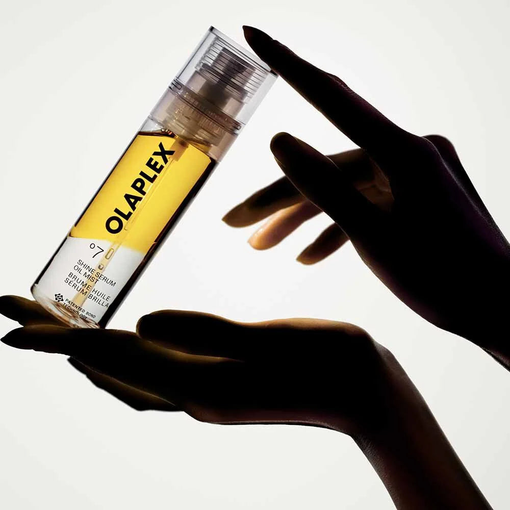 A clear cylindrical bottle of OLAPLEX hair product with a yellow label and transparent cap, held between two dark silhouetted hands against a bright white background.