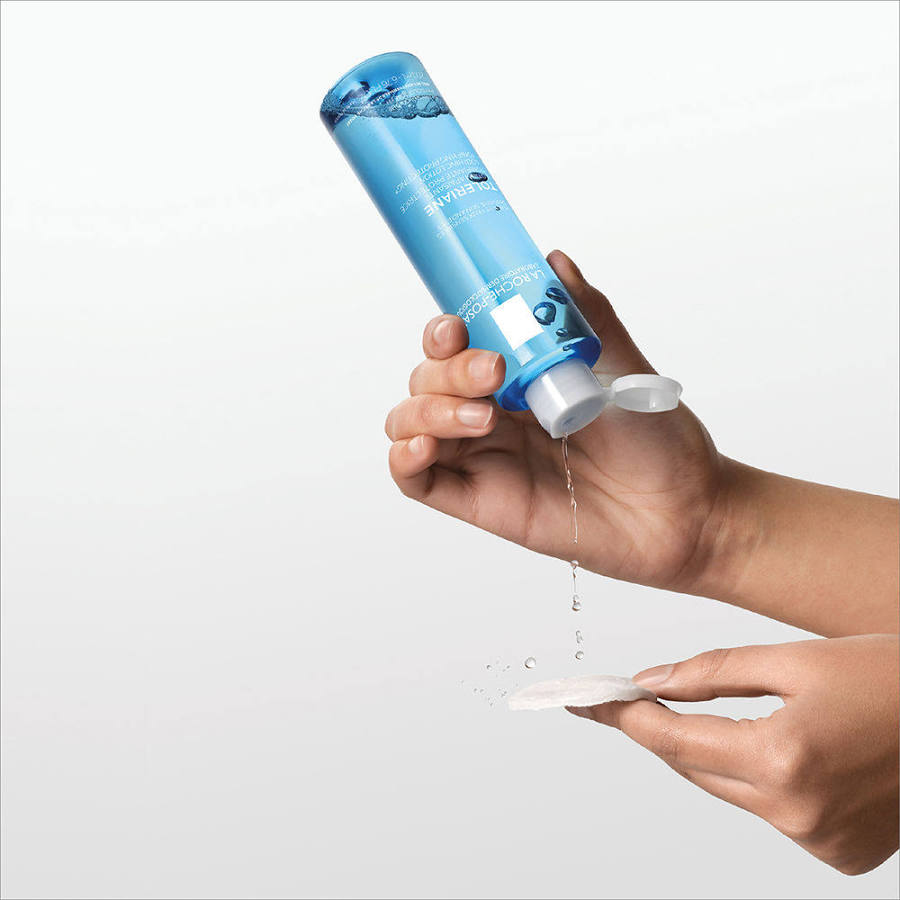 Hand holding a blue bottle with water being poured onto a white surface against a light gray background
