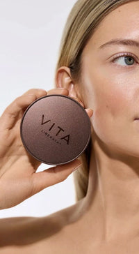 Person holding a round brown compact with 'Vita' branding against a neutral background
