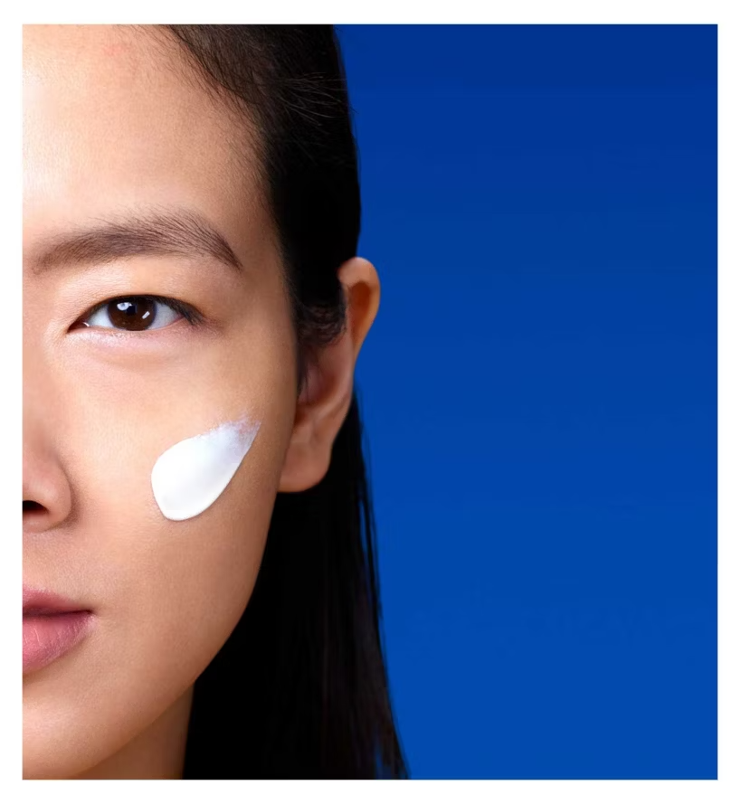 Close-up of a woman's face with La Roche Posay Anthelios Age Correct SPF product application on her cheek against a blue background