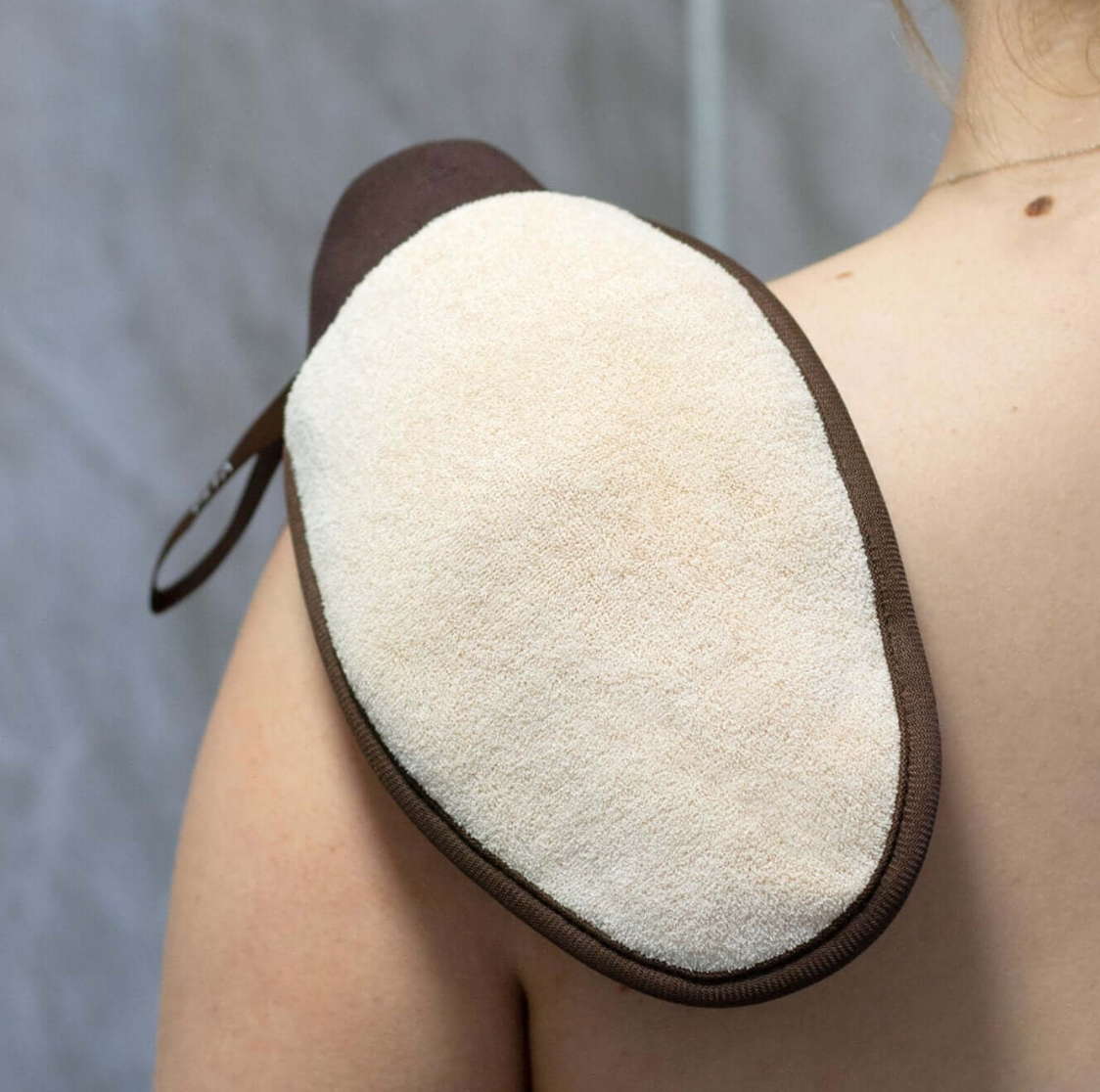 A person using a Vita Liberata Dual-sided exfoliating mitt on their back, with the mitt showing a beige, textured surface suitable for skin exfoliation.