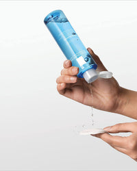 Hand holding a blue bottle with water being poured onto a white surface against a light gray background